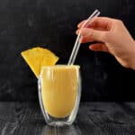 A side view of a hand holding a glass straw in a clear glass filled with spicy pineapple turmeric smoothie.