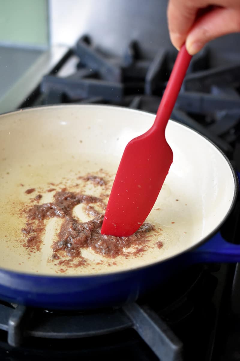 A red spatula is breaking up anchovy fillets in a ceramic cast iron skillet.