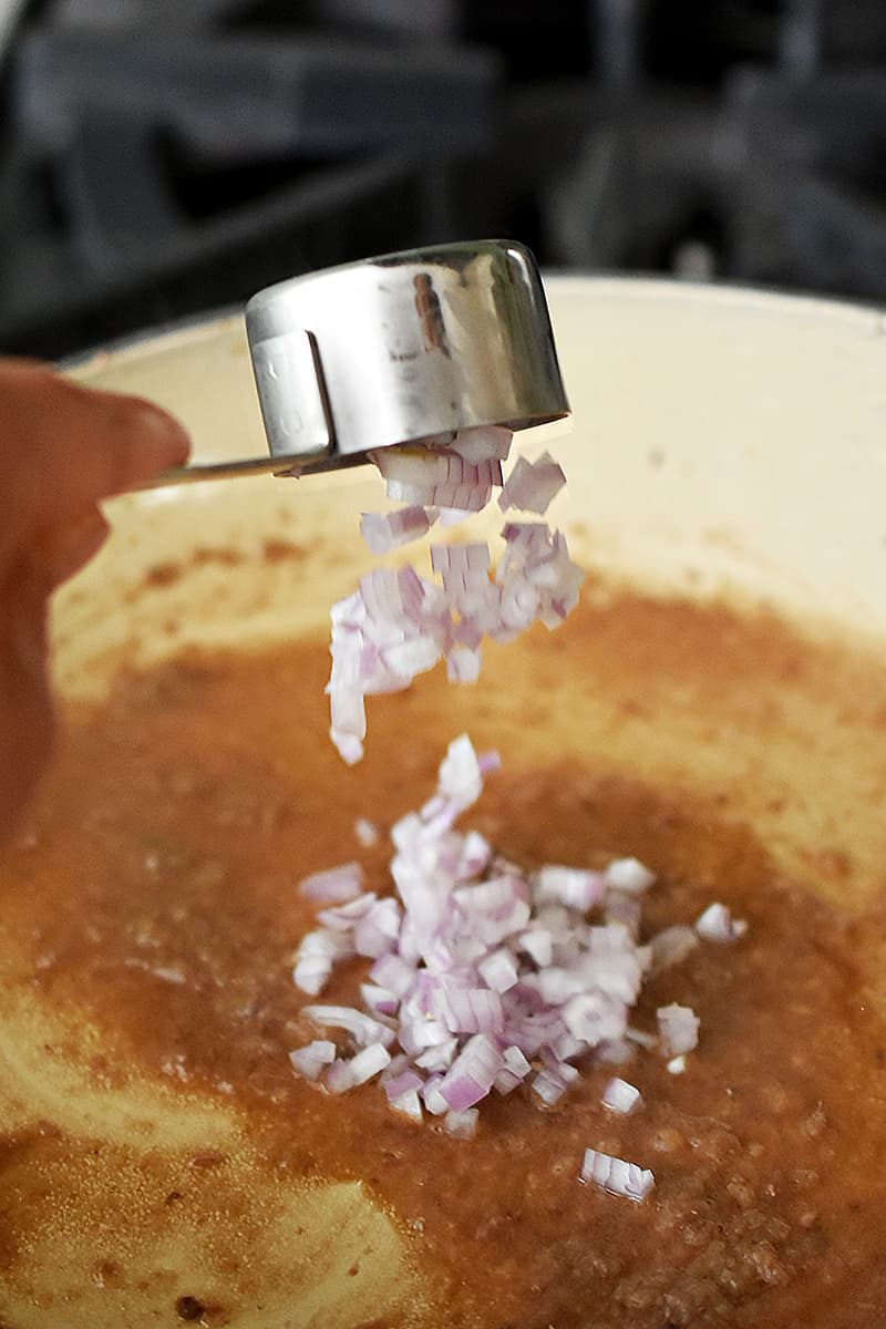 A hand is adding a measuring cup filled with minced shallots into a skillet to make garlic anchovy vinaigrette
