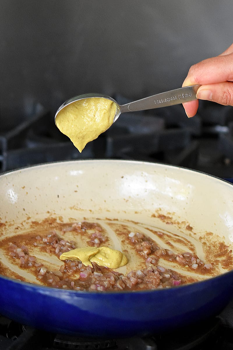 A hand is adding a measuring spoon filled with Dijon mustard into a skillet filled with garlic anchovy vinaigrette.