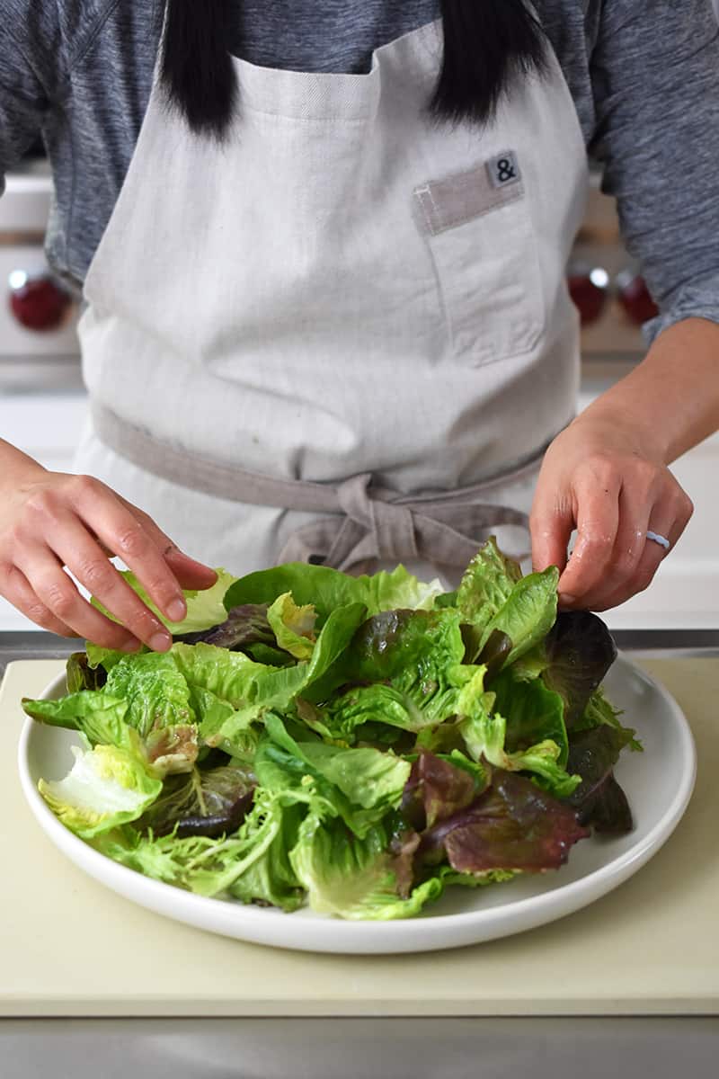 A person in a white apron is arranging mixed baby lettuce on a platter.