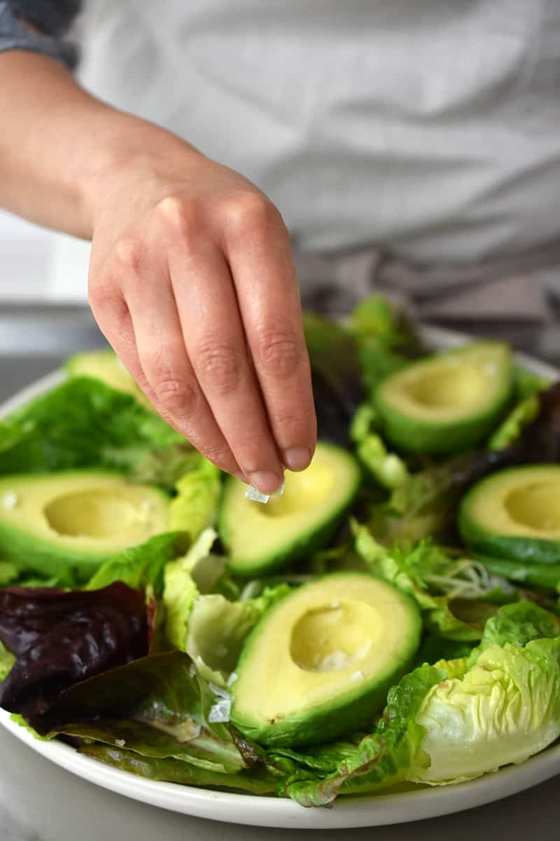 A person in a white apron is sprinkling salt on an avocado salad