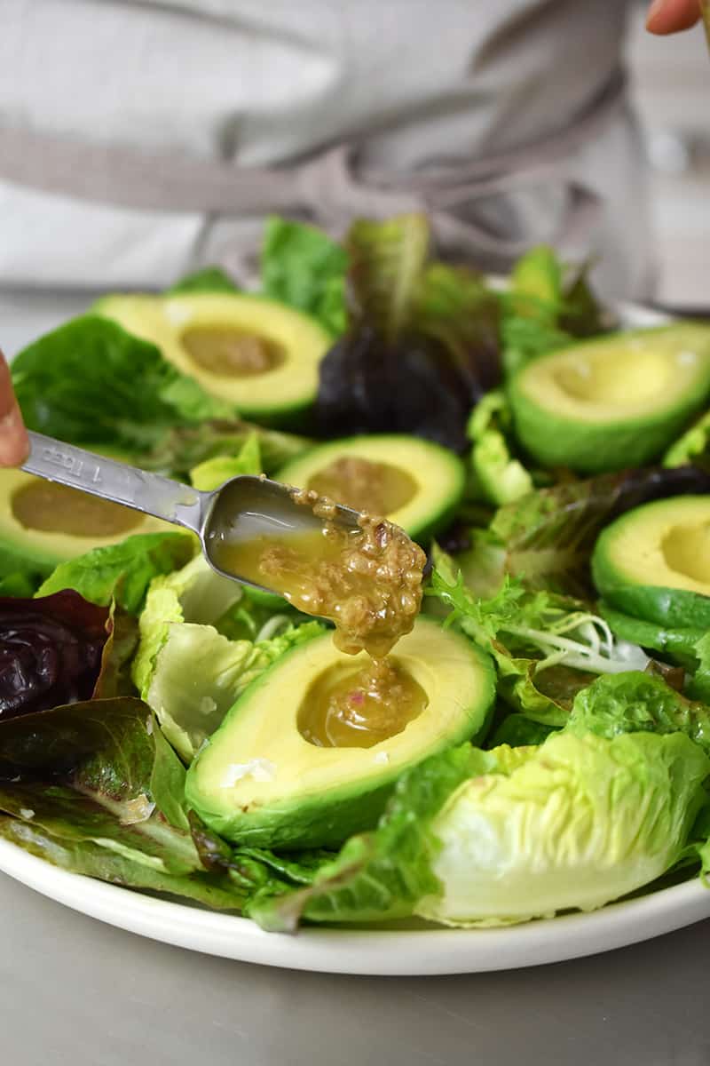 Adding a spoonful of garlic anchovy vinaigrette into each halved avocado on the salad greens.