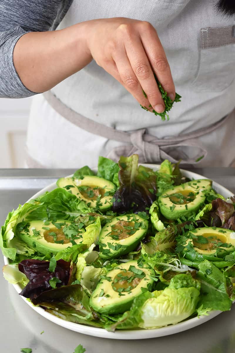 A person in a white apron is sprinkling fresh herbs on the avocado salad.