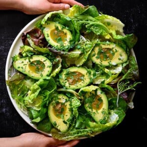 An overhead shot of two hands holding a round platter filled with avocado salad and garlic anchovy vinaigrette