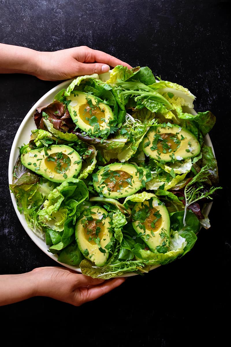 An overhead shot of two hands holding a round platter filled with avocado salad and garlic anchovy vinaigrette 