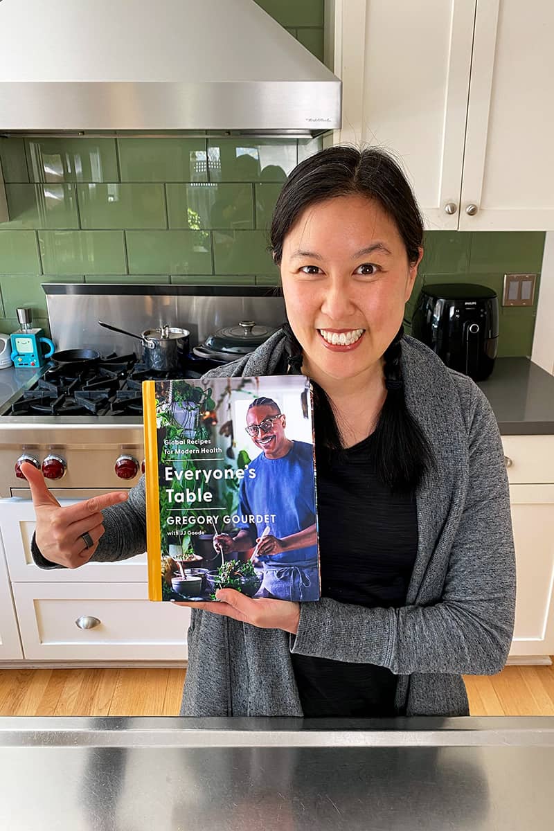 A smiling Asian woman in a kitchen is holding up a copy of Gregory Gourdet’s Everyone's Table cookbook