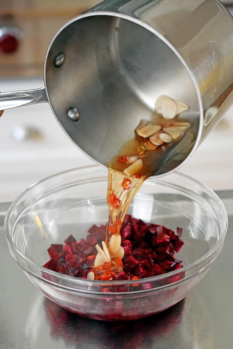 A saucepan filled with hot vinegar, garlic, and Thai chili peppers is being poured into a glass bowl with diced beets.