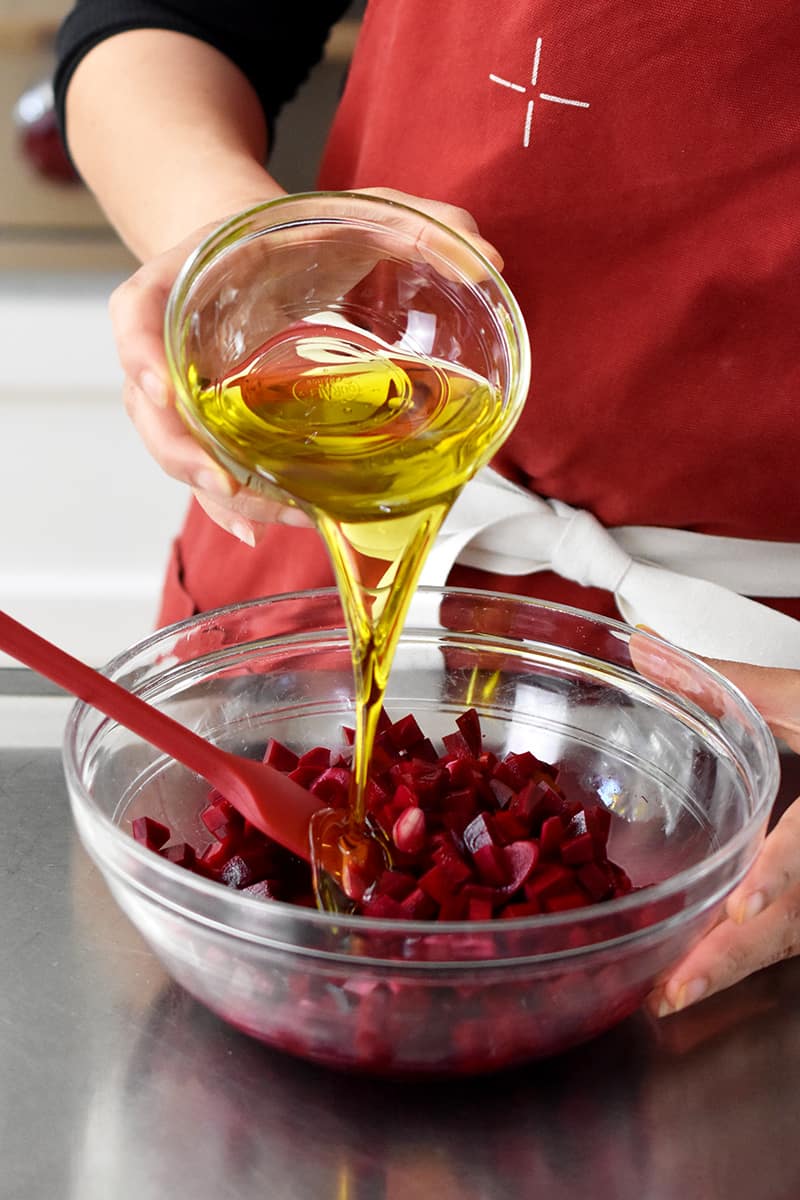 Someone in a red apron is pouring a small glass bowl filled with extra virgin olive oil into a glass bowl filled with diced beets and vinegar.