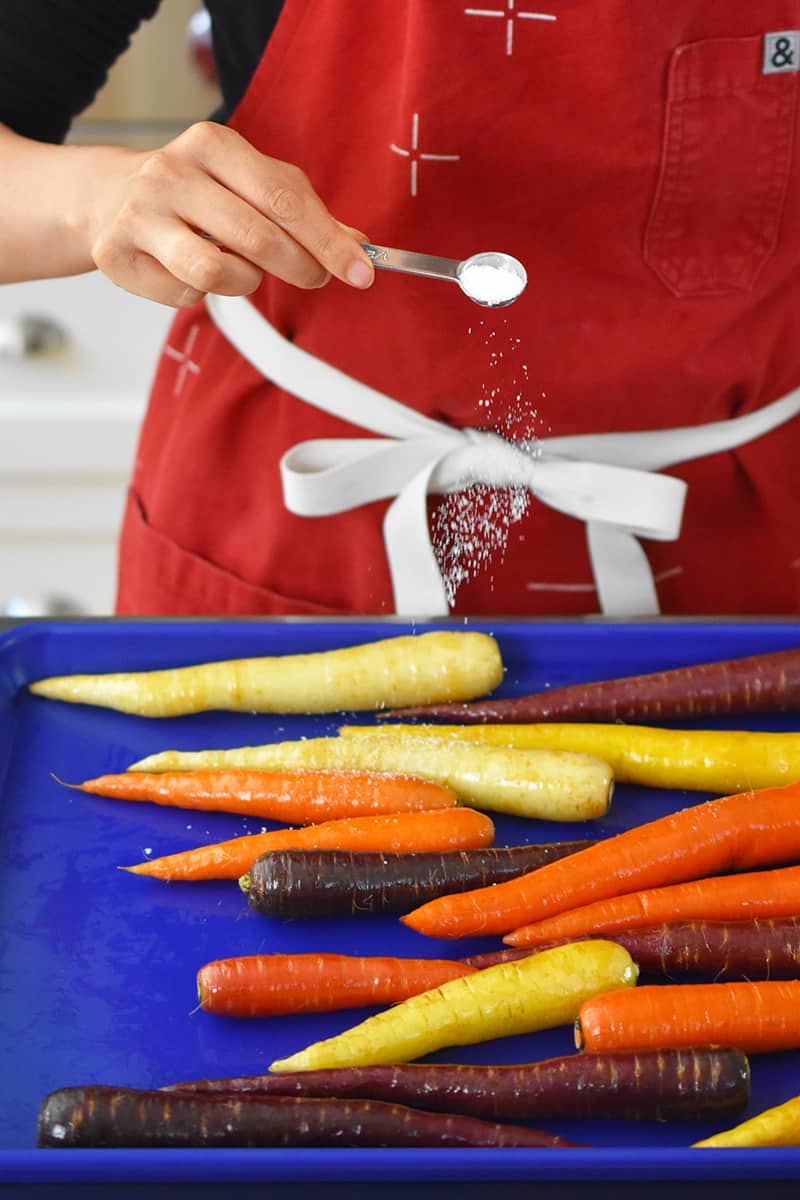 A person in a red apron is sprinkling salt on a blue rimmed baking sheet filled with colorful carrots