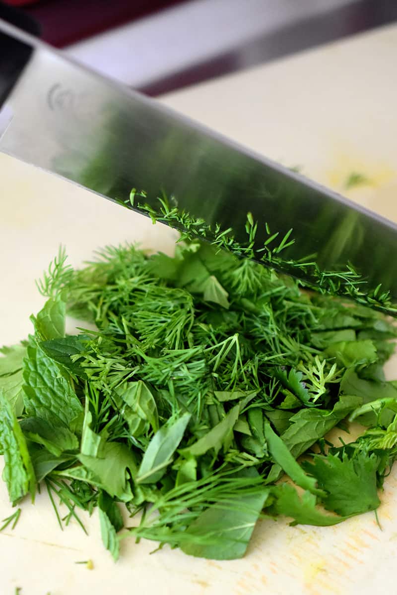 An overhead shot of a chef's knife cutting a mixture of herbs on a beige cutting board.