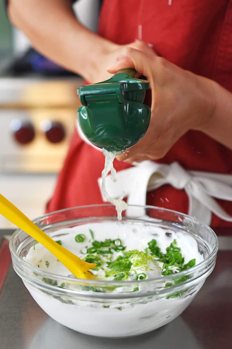 Someone in a red apron is using a green lime juicer to squeeze lime juice into a bowl filled with coconut yogurt and fresh herbs.