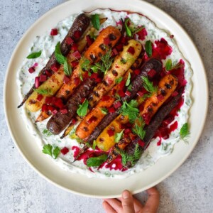 A hand is holding a round platter filled with grilled carrots with herby coconut yogurt and spicy beet vinaigrette