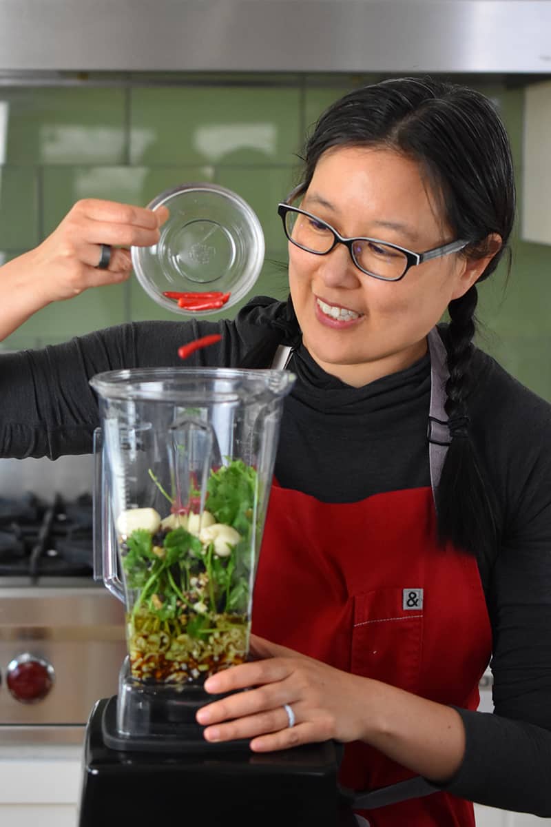 A smiling Asian woman is adding Thai Chili peppers to a blender to make a grilled chicken marinade.