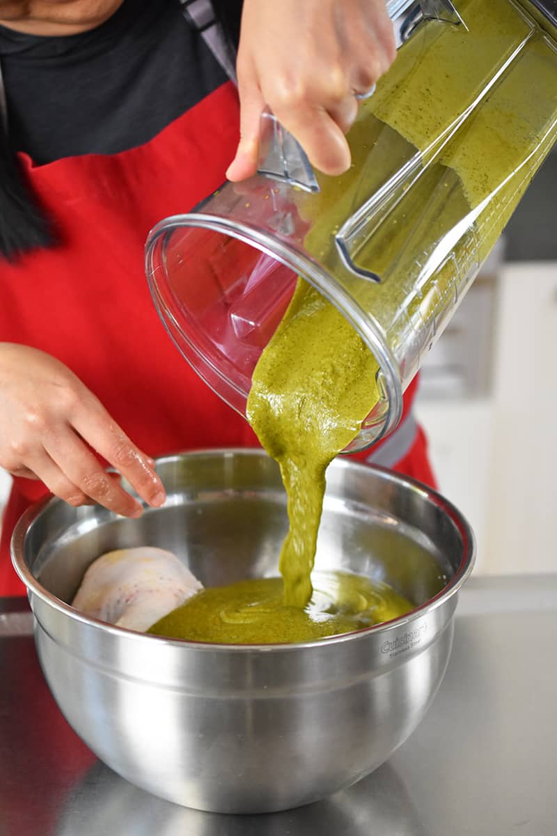 A person in a red apron is pouring a greenish brown marinade into a silver mixing bowl filled with salted chicken thighs.