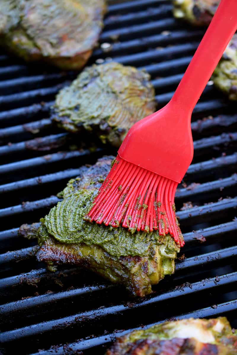 A red silicone brush is adding marinade to the skin side of grilled chicken thighs. 