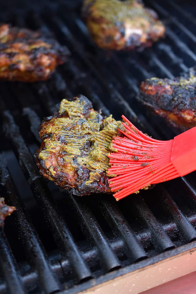 A silicone brush is brushing marinade on the meat side of a grilled chicken thigh on a gas grill.