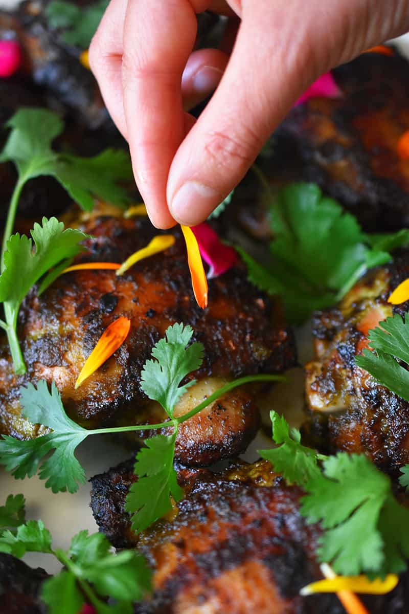A hand is adding edible flower petals to a platter of grilled chicken with fresh cilantro.