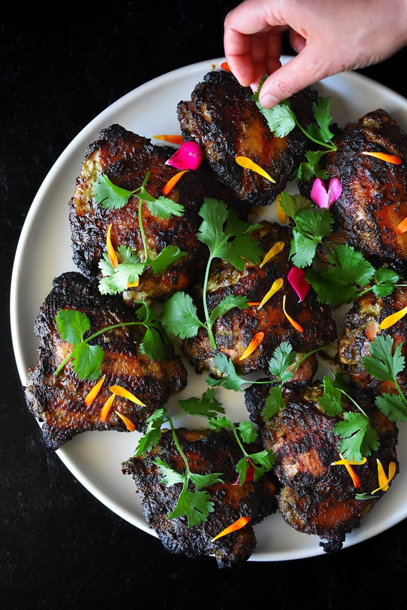 A hand is adding fresh cilantro and edible flower petals to a platter of grilled chicken.