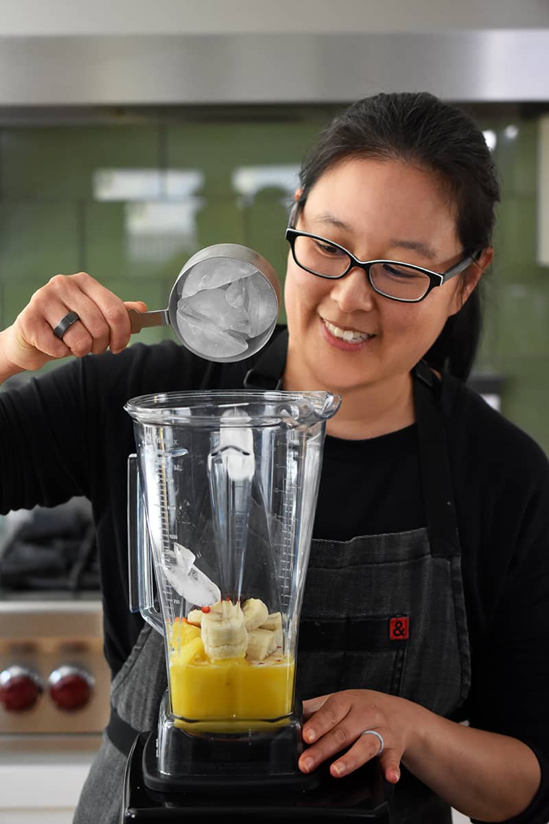 A smiling Asian woman is adding a cup filled with ice cubes to a Vitamix blender filled with pineapple smoothie ingredients.