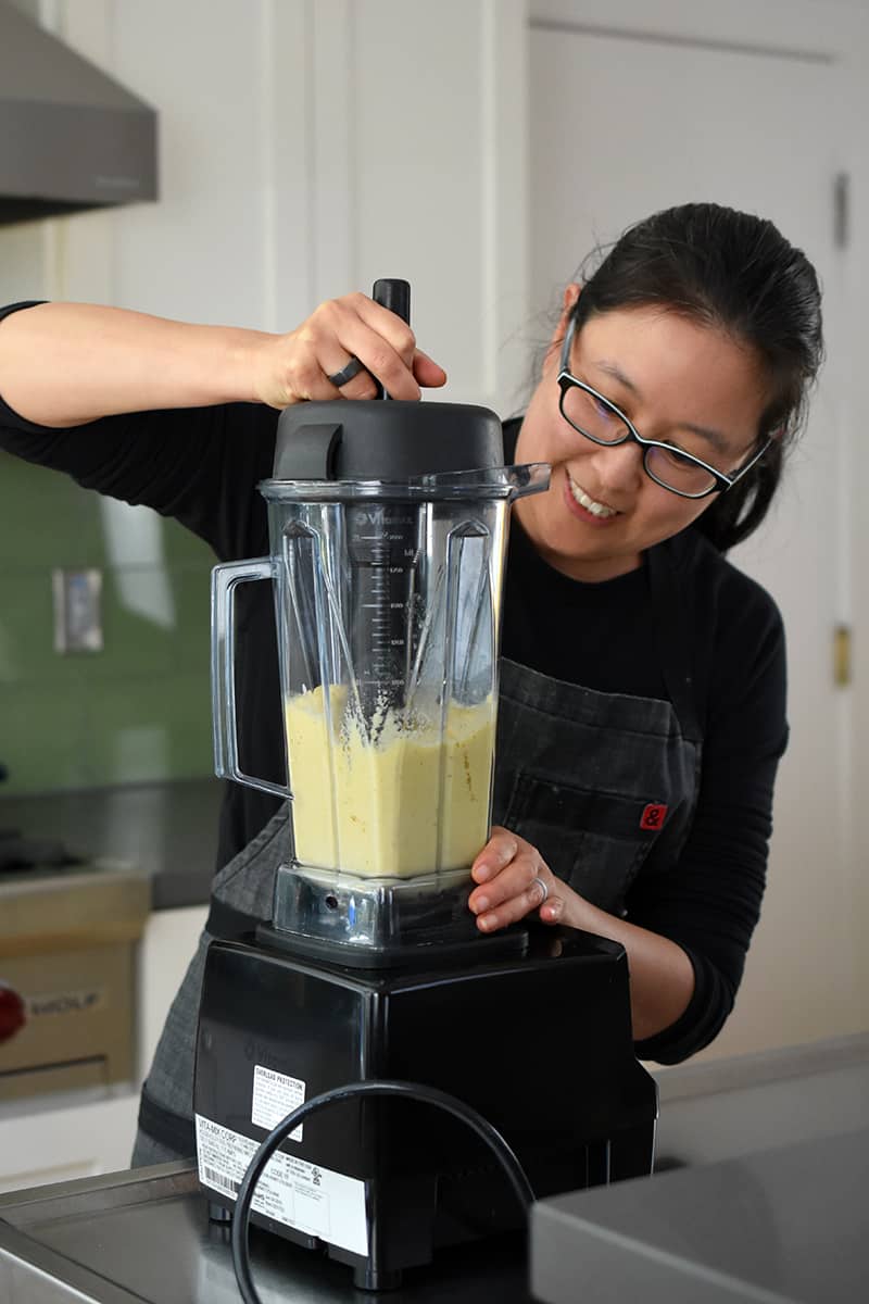 A smiling Asian woman is blending a pineapple smoothie in a Vitamix.