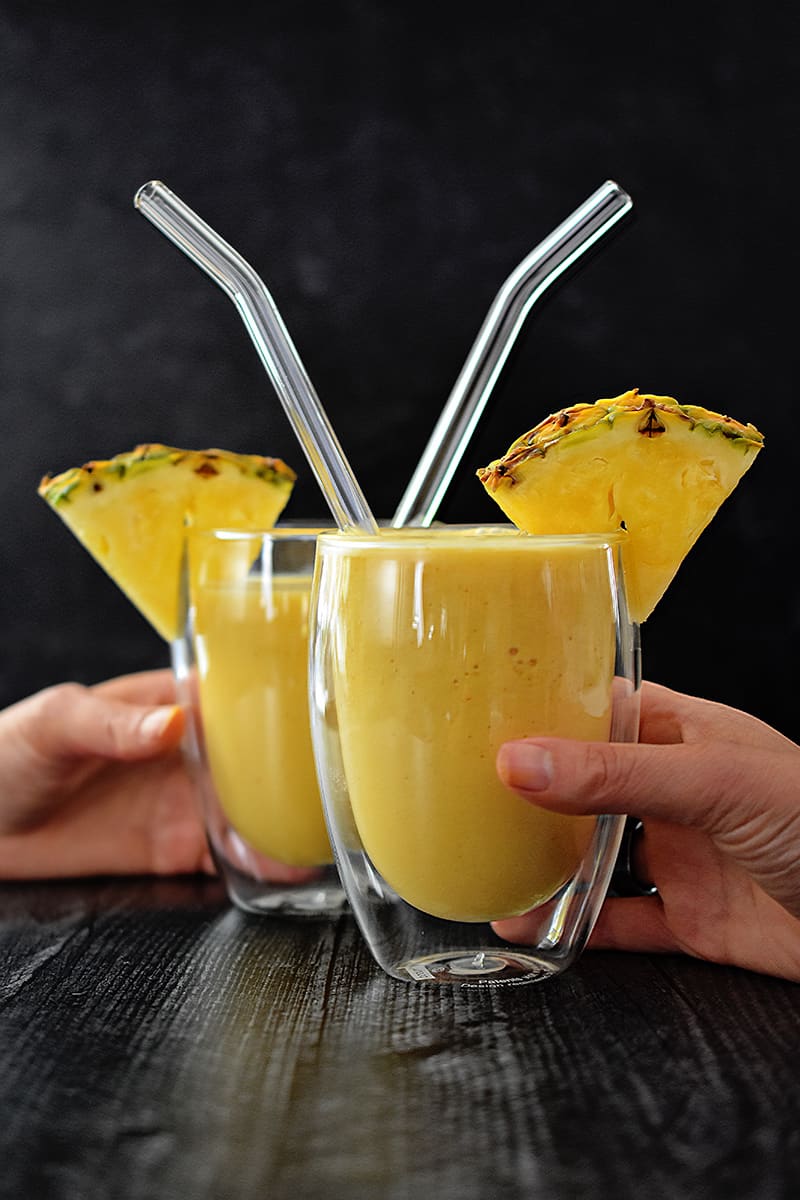 Two glasses of pineapple smoothie in clear glasses are being held by hands. There is a black backdrop and tabletop.