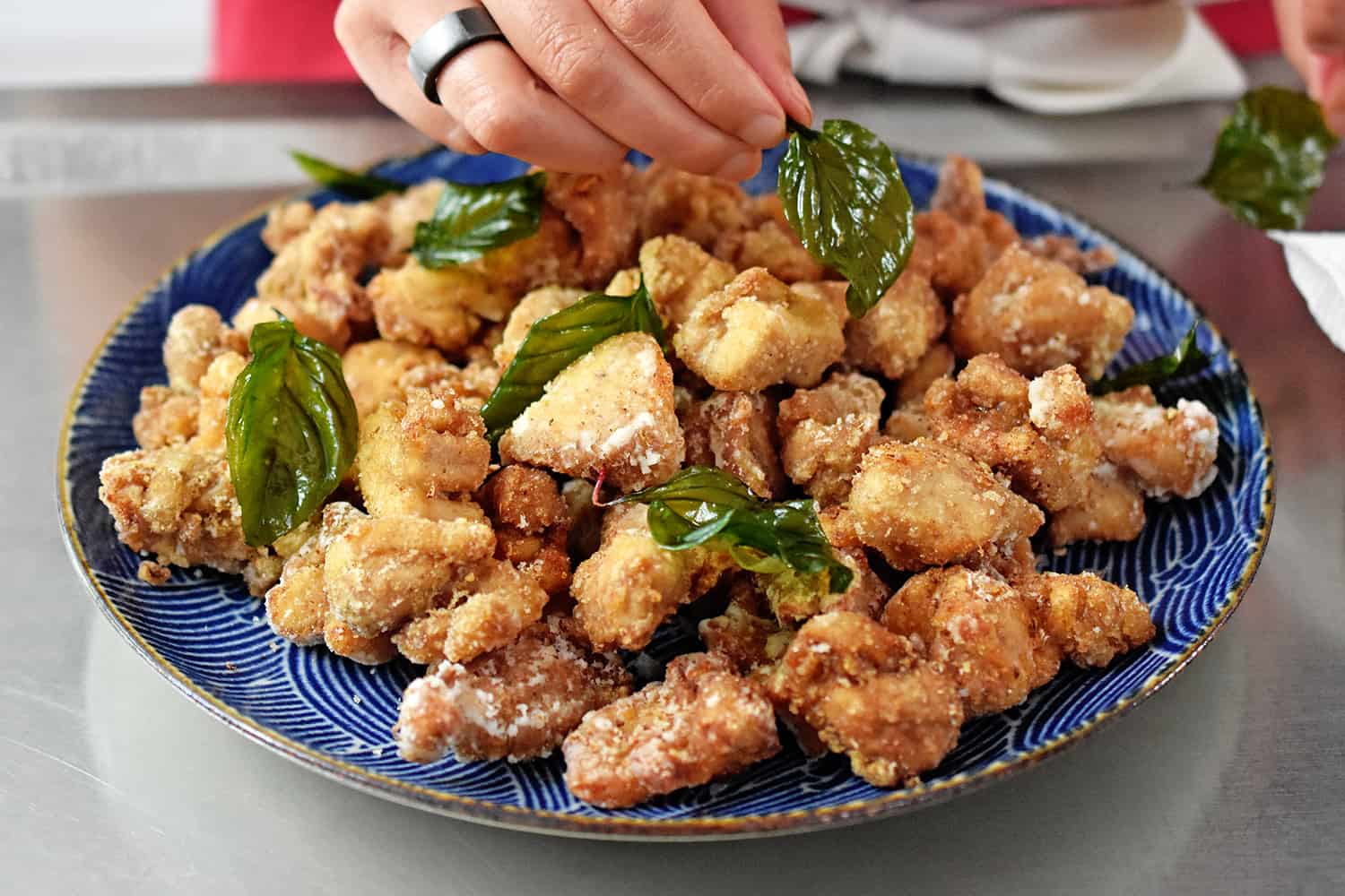 A person is adding fried Thai basil leaves to a platter of deep fried Taiwanese popcorn chicken