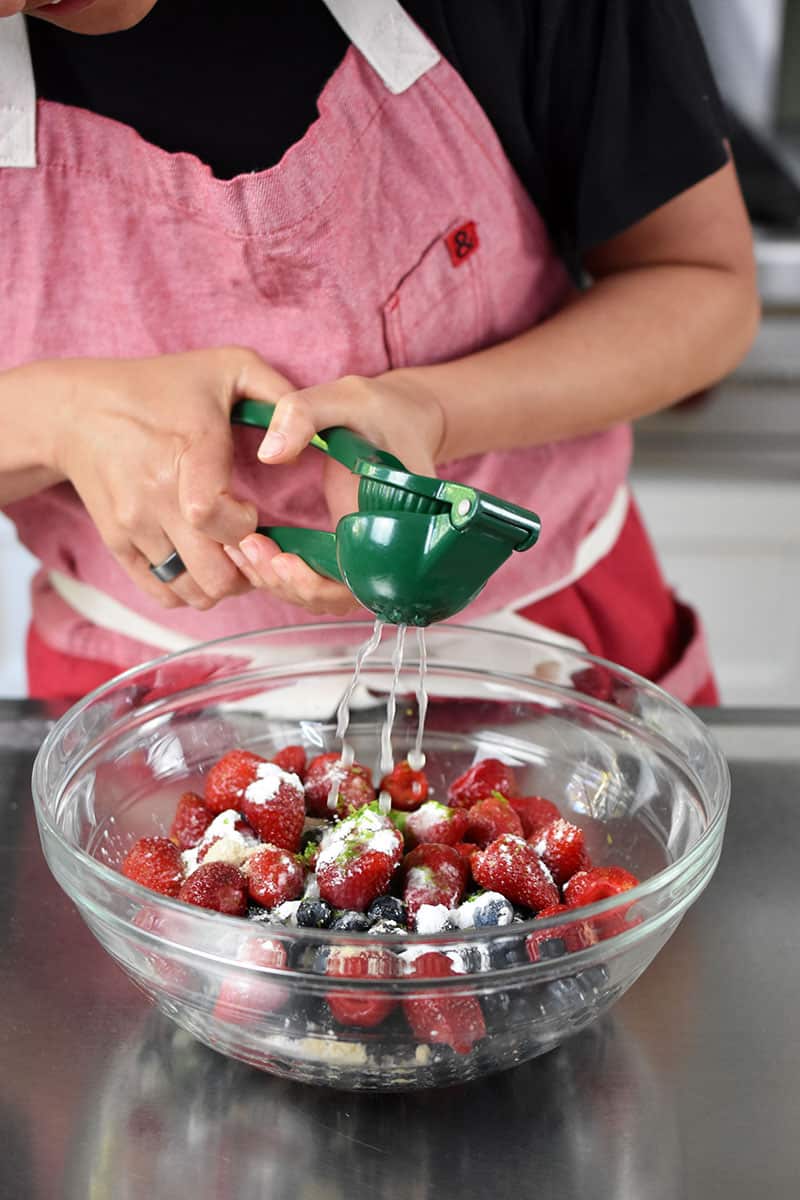 A person in a red apron is adding lime juice to a bowl filled with mixed berries