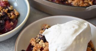 A shallow bowl contains a serving of paleo berry crisp topped with whipped coconut cream. It is in front of a baking dish filled with the berry crisp fresh out of the oven.