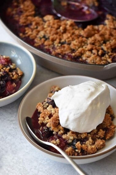 A shallow bowl contains a serving of paleo berry crisp topped with whipped coconut cream. It is in front of a baking dish filled with the berry crisp fresh out of the oven.