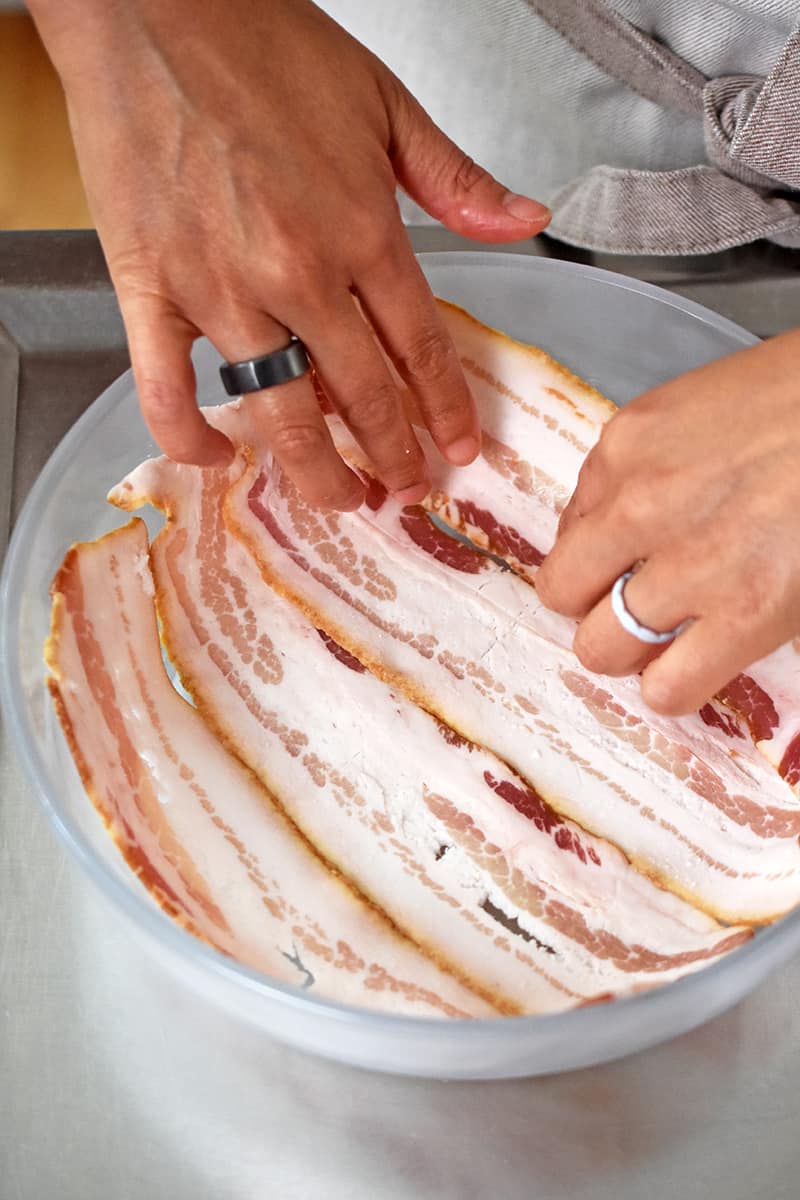 A hand is laying four slices of bacon in a single layer in a flat glass microwave safe container