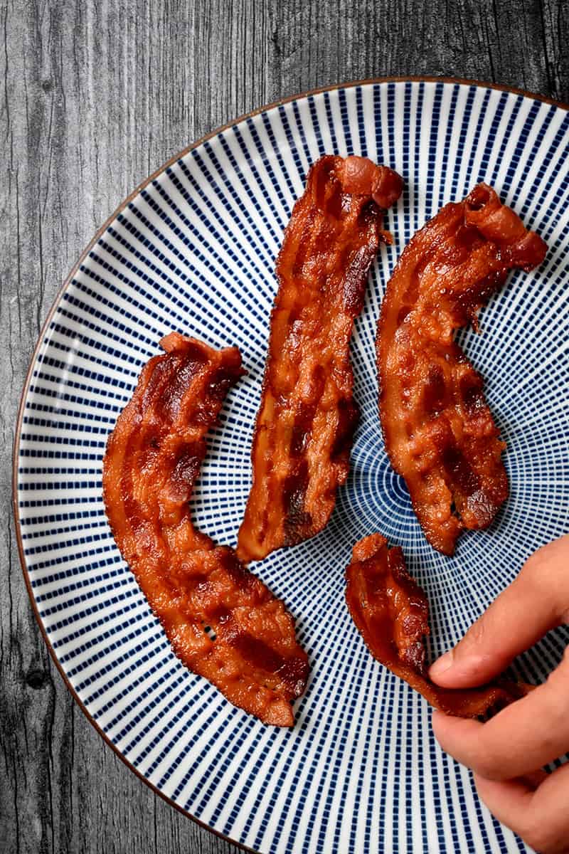 A hand grabbing a piece of crispy microwave bacon from a blue and white plate
