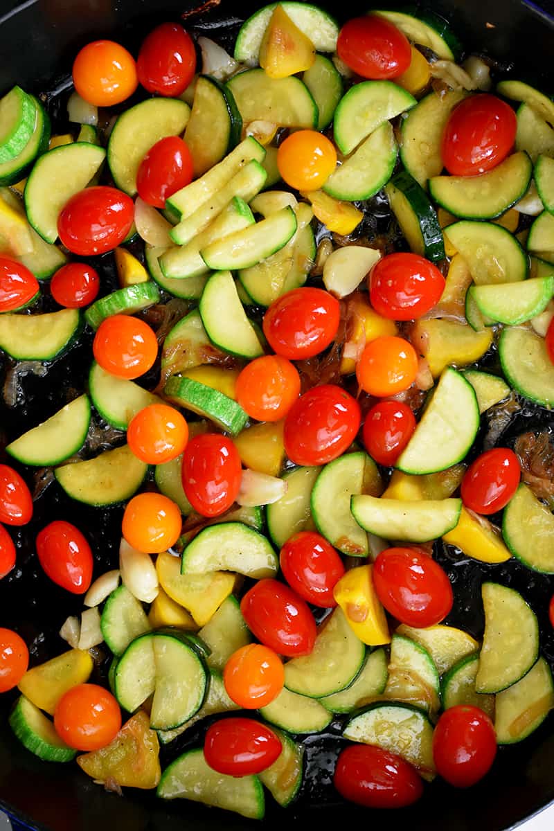An overhead shot of a skillet filled with sautéed sliced summer squash, browned shallot slices, and cherry tomatoes