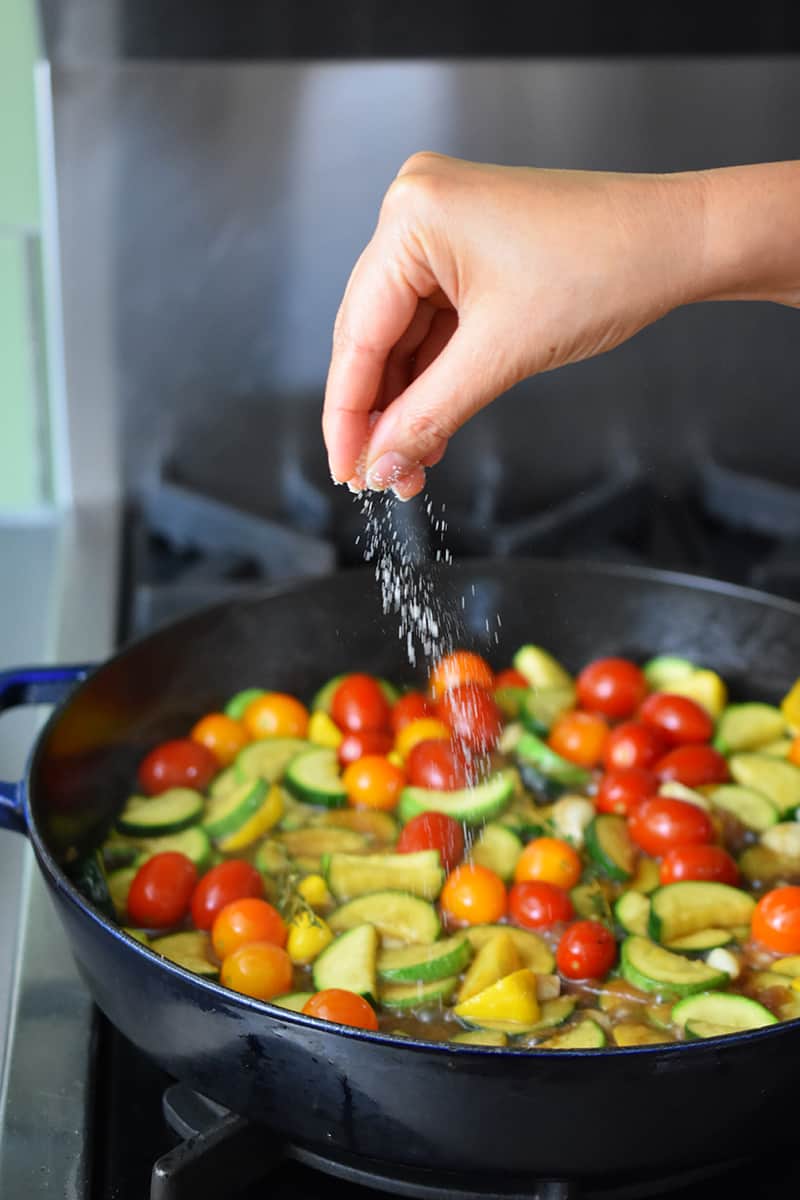 A hand is adding salt to a skillet filled with sliced summer squash, cherry tomatoes, garlic, shallot, and thyme.