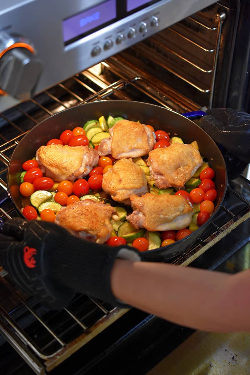 Someone placing a skillet with crispy chicken thighs and summer vegetables into an open oven