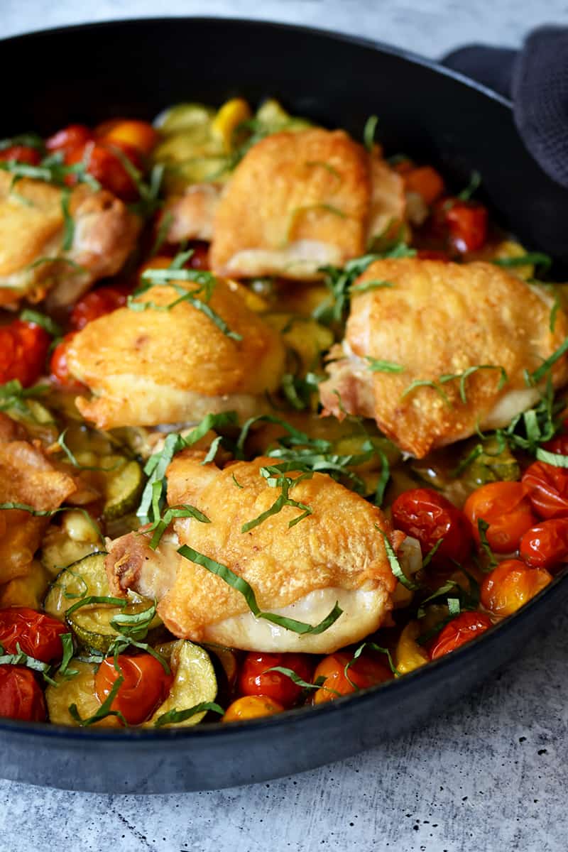 A side view of a skillet filled with crispy baked chicken thighs and zucchini, cherry tomatoes, and fresh basil.