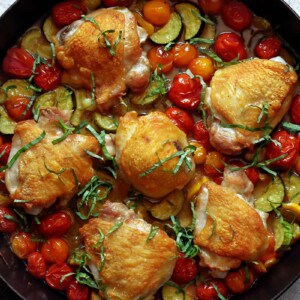 An overhead shot of a skillet filled with crispy chicken thighs, cherry tomatoes, zucchini, and basil