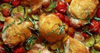 An overhead shot of a skillet filled with crispy chicken thighs, cherry tomatoes, zucchini, and basil