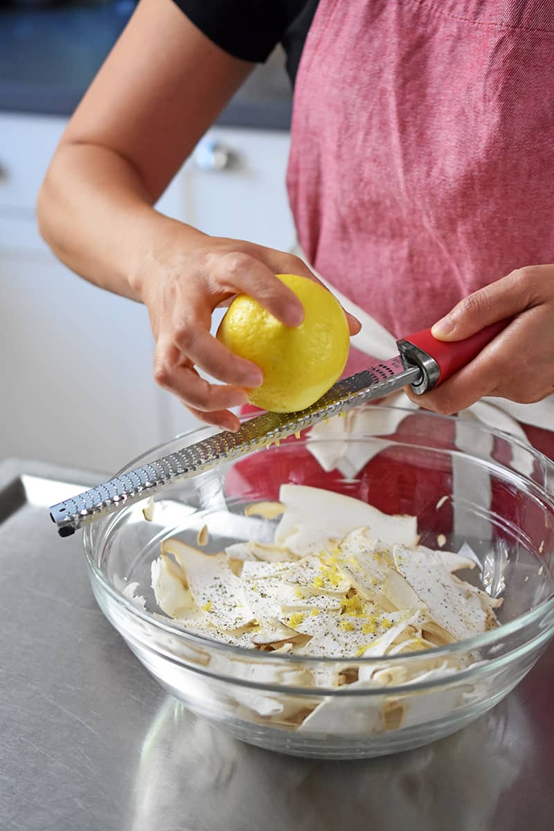 A person in a red apron is grating fresh lemon zest on a mushroom salad