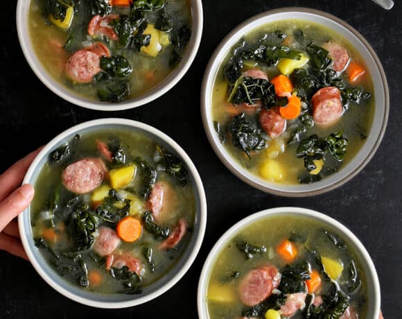 An overhead shot of someone grabbing one of four bowls filled with sausage kale soup.