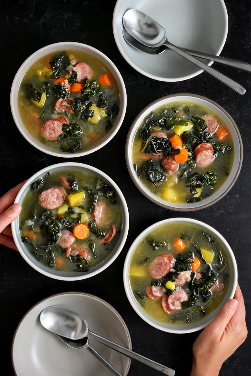 An overhead shot of someone grabbing one of four bowls filled with sausage kale soup.