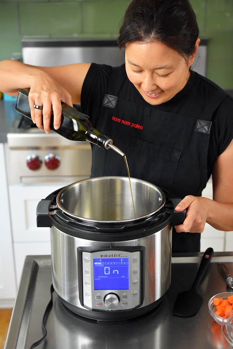 A smiling Asian woman is adding extra virgin olive oil to an Instant Pot on the sauté function
