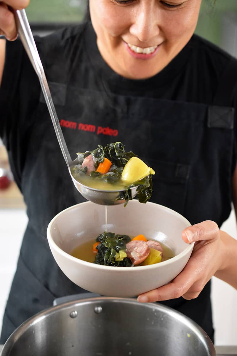 A smiling Asian woman is ladling a bowl of Instant Pot kale and sausage soup