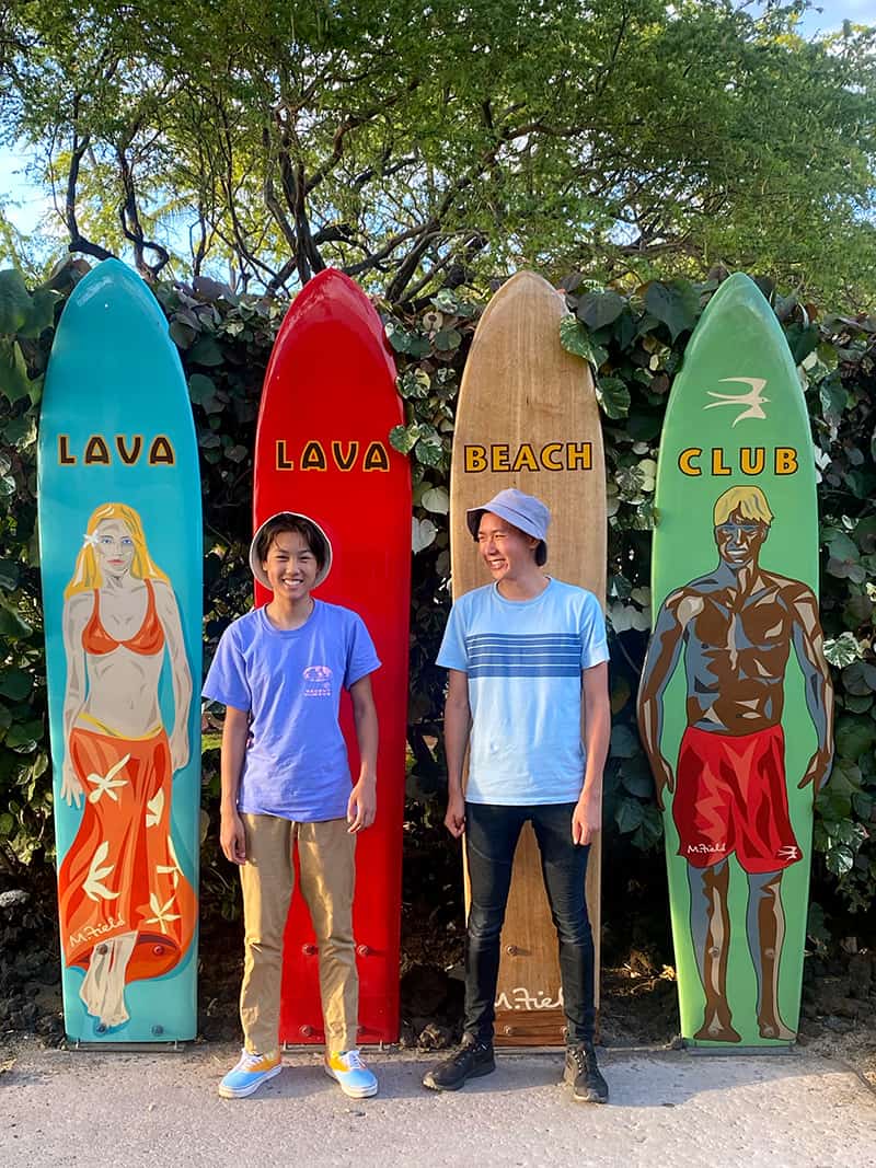 Two Asian teenage boys are posed in front of surf boards that say Lava Lava Beach Club