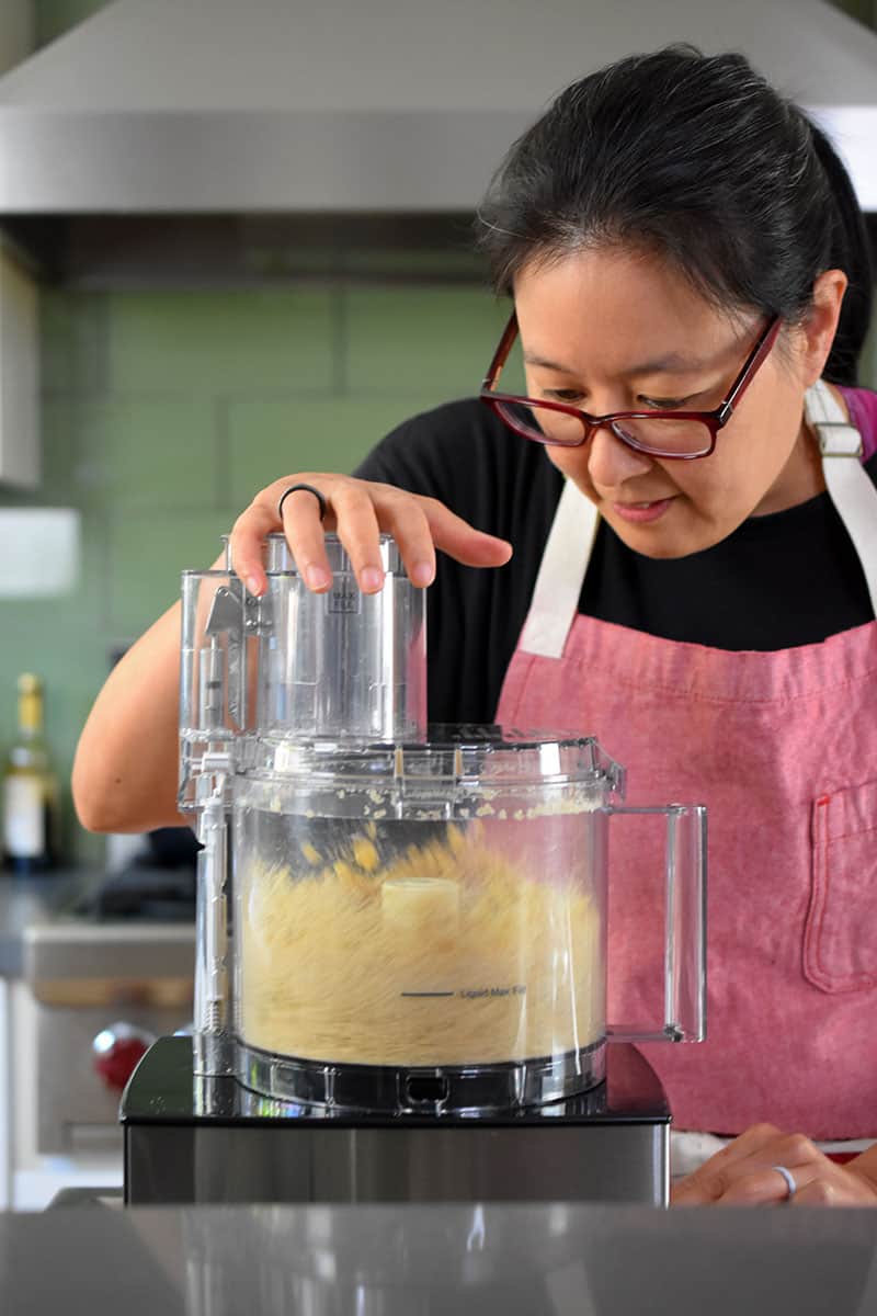 An Asian woman in a red apron is pulsing the topping ingredients for paleo apple crisp in a food processor.