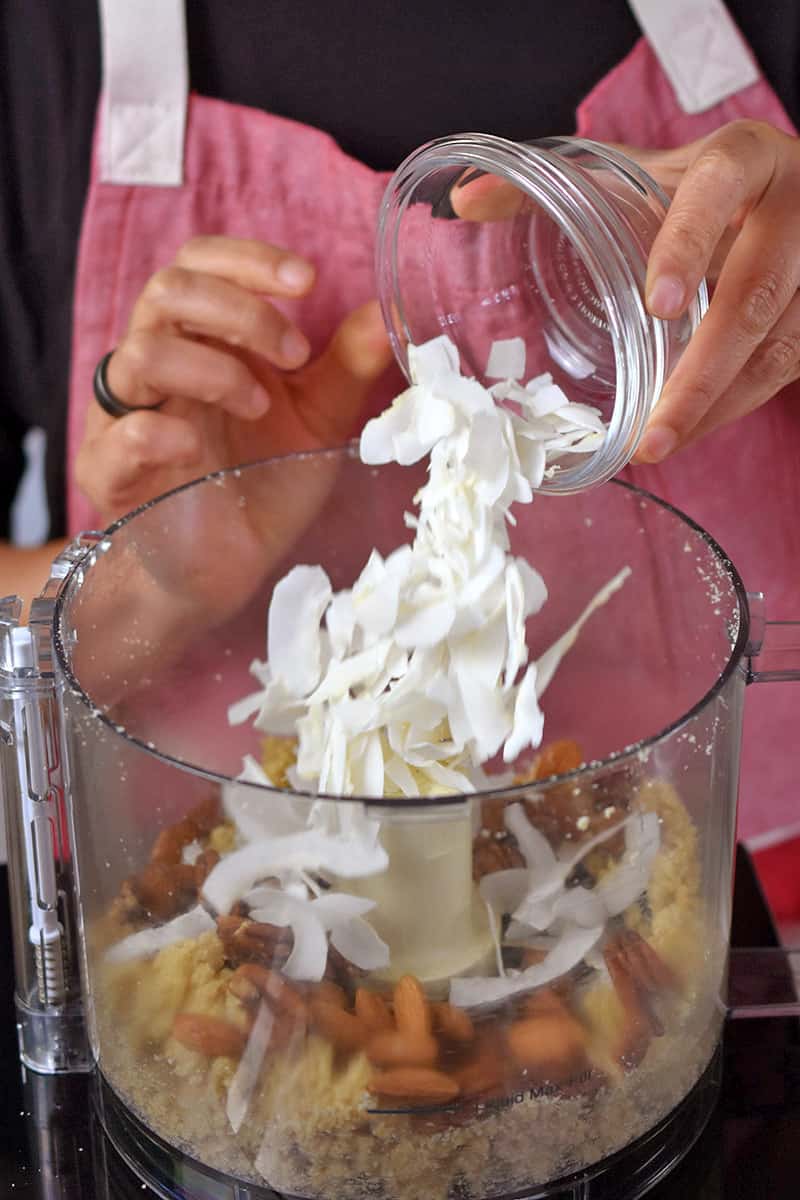 A person in a red apron is adding coconut flakes to a food processor filled with paleo apple crisp topping ingredients.