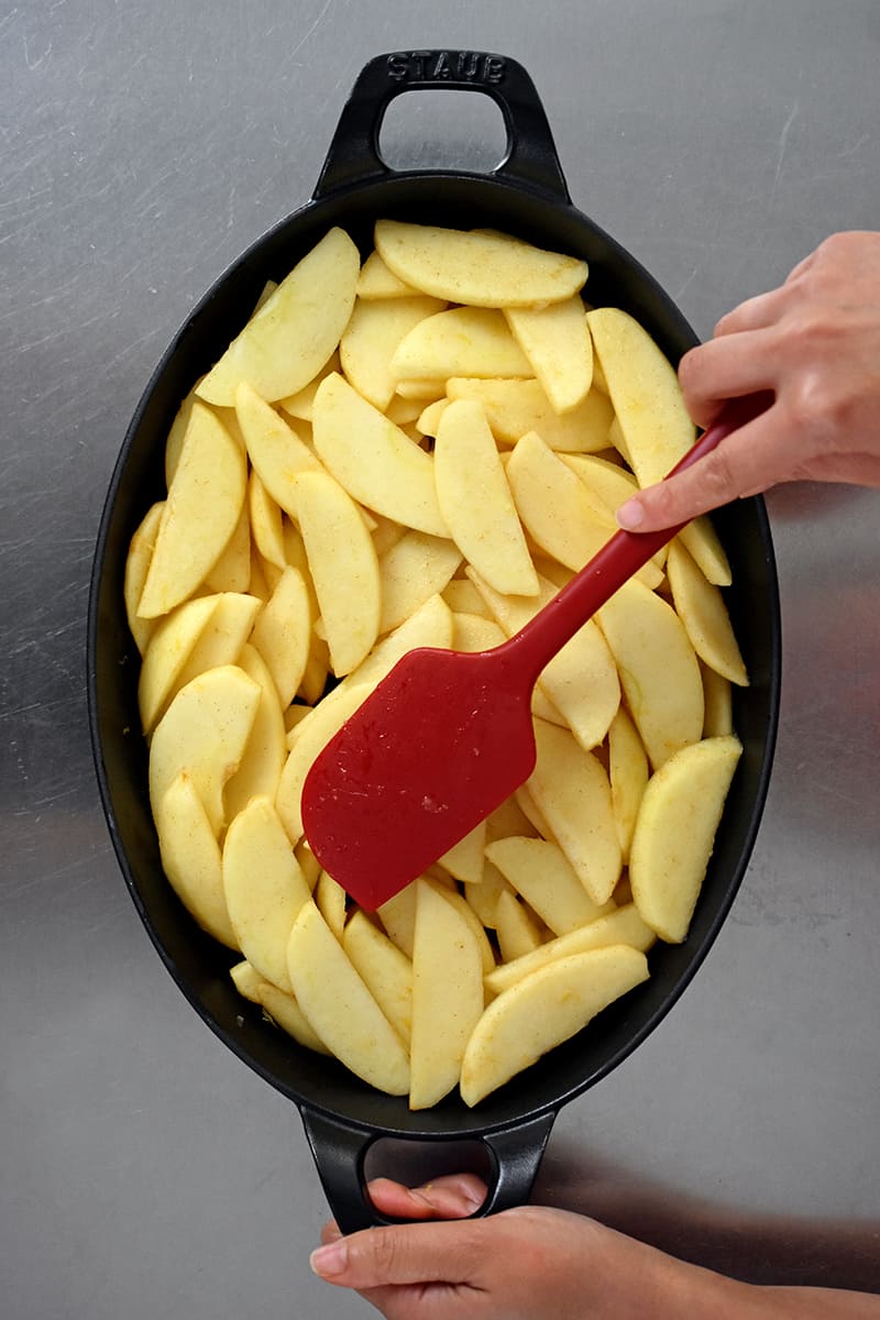 An overhead shot of an oval baking dish filled with sliced apples. A red silicone spatula is making an even layer.