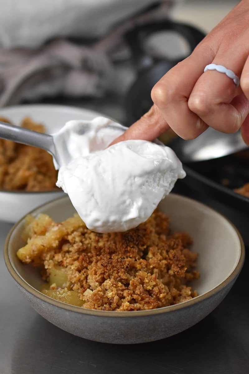 Topping a shallow bowl filled with paleo apple crisp with whipped coconut cream.
