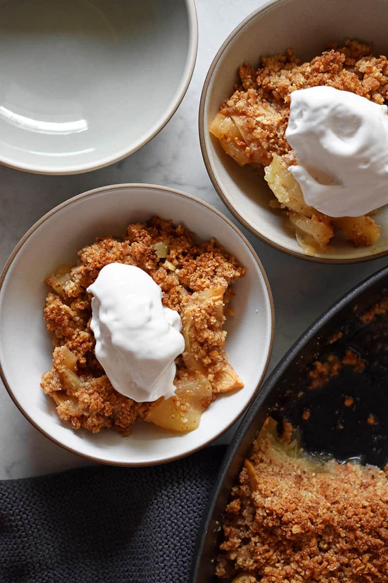 An overhead shot of two bowls of paleo, vegan, and gluten-free apple crisp topped with dollops of whipped coconut cream.