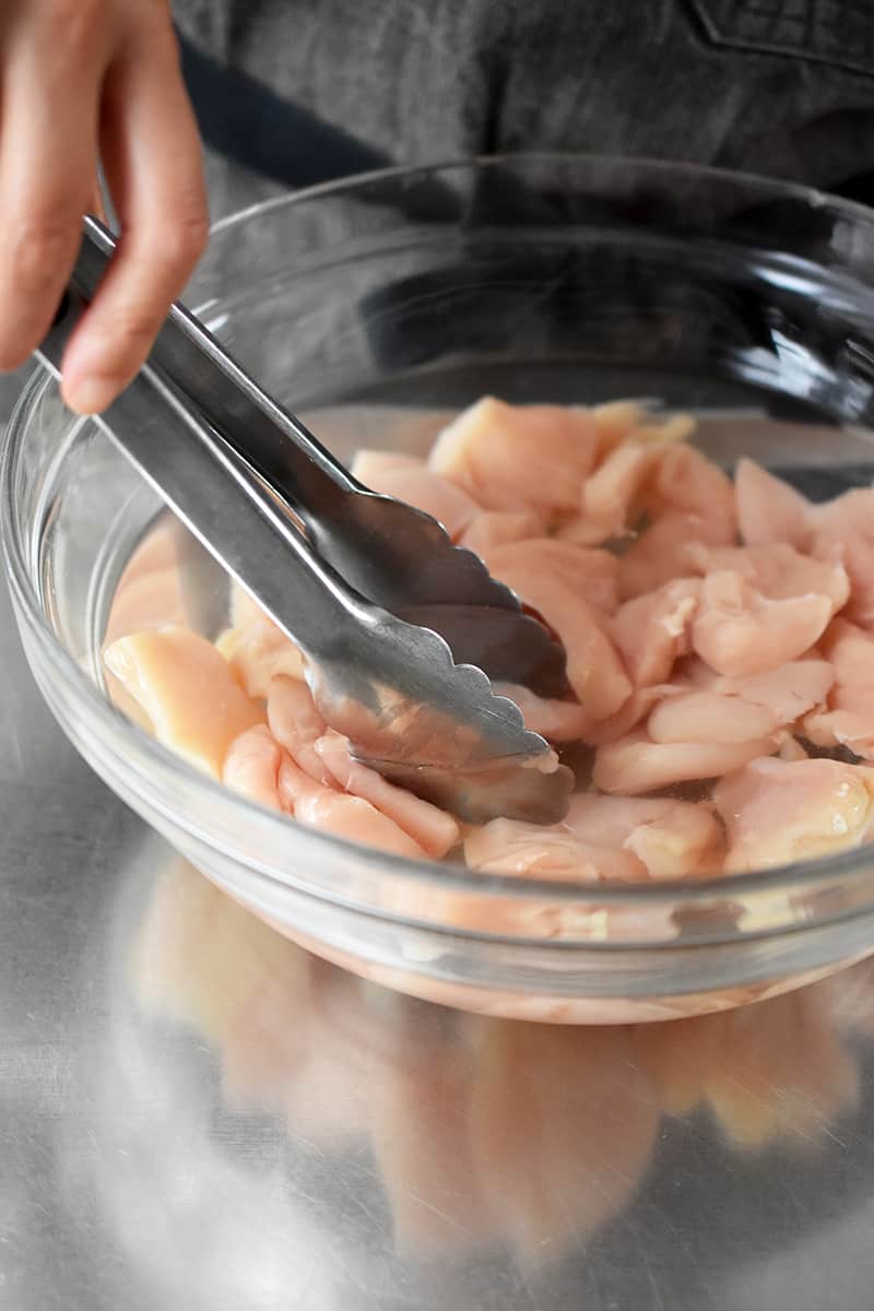 Using tongs to submerge the chicken breast pieces in a salt brine.